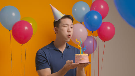 Studio-Portrait-Of-Man-Wearing-Party-Hat-Celebrating-Birthday-Blowing-Out-Candles-On-Cake